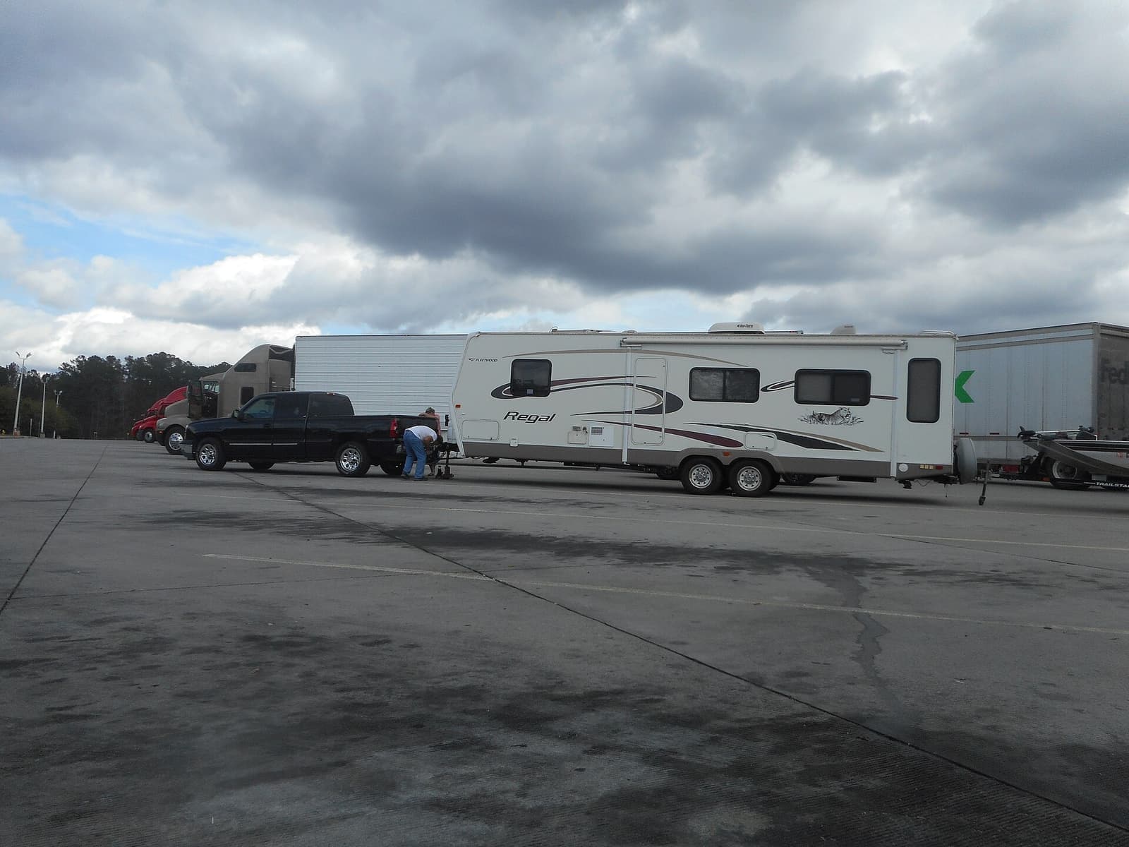 A pickup truck hitched to a travel trailer at a highway rest area, with a person working at the coupler